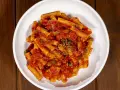 Un piatto di pasta alla pizzaiola, con rigatoni conditi da un sugo rosso ricco di pomodoro, capperi e origano, servito in una ciotola bianca su un tavolo di legno.