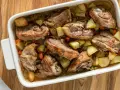 Pezzi succulenti di capretto al forno alla siciliana con patate e carote in una teglia bianca.