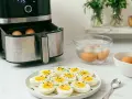Uova sode in friggitrice ad aria pronte per essere gustate. Alcune ancora nella friggitrice, altre in acqua ghiacciata, e un piatto pieno di uova sode tagliate a metà, condite con erba cipollina e pepe.