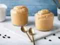 Due bicchieri di cremosa schiuma di caffè, perfetti per una ricetta crema di caffè senza panna. Due cucchiaini dorati e chicchi di caffè completano la scena.