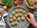 Polpette di pollo pronte per essere cucinate, disposte su un piatto grigio, con ingredienti freschi come cipolle e pomodoro sullo sfondo.
