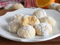 Biscotti alla ricotta soffici e profumati, cosparsi di zucchero a velo, con una fetta d'arancia sullo sfondo.
