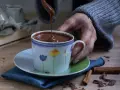 Una mano versa cioccolata fatta in casa in una tazza fiorata. Profumo di cannella e scaglie di cioccolato sul tavolo.