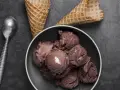 Coppa di **gelato al cioccolato** cremosa, con coni vuoti e un cucchiaio d'argento su sfondo scuro.