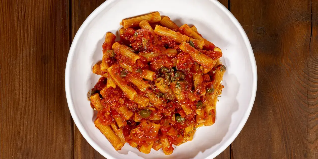 Un piatto di pasta alla pizzaiola, con rigatoni conditi da un sugo rosso ricco di pomodoro, capperi e origano, servito in una ciotola bianca su un tavolo di legno.