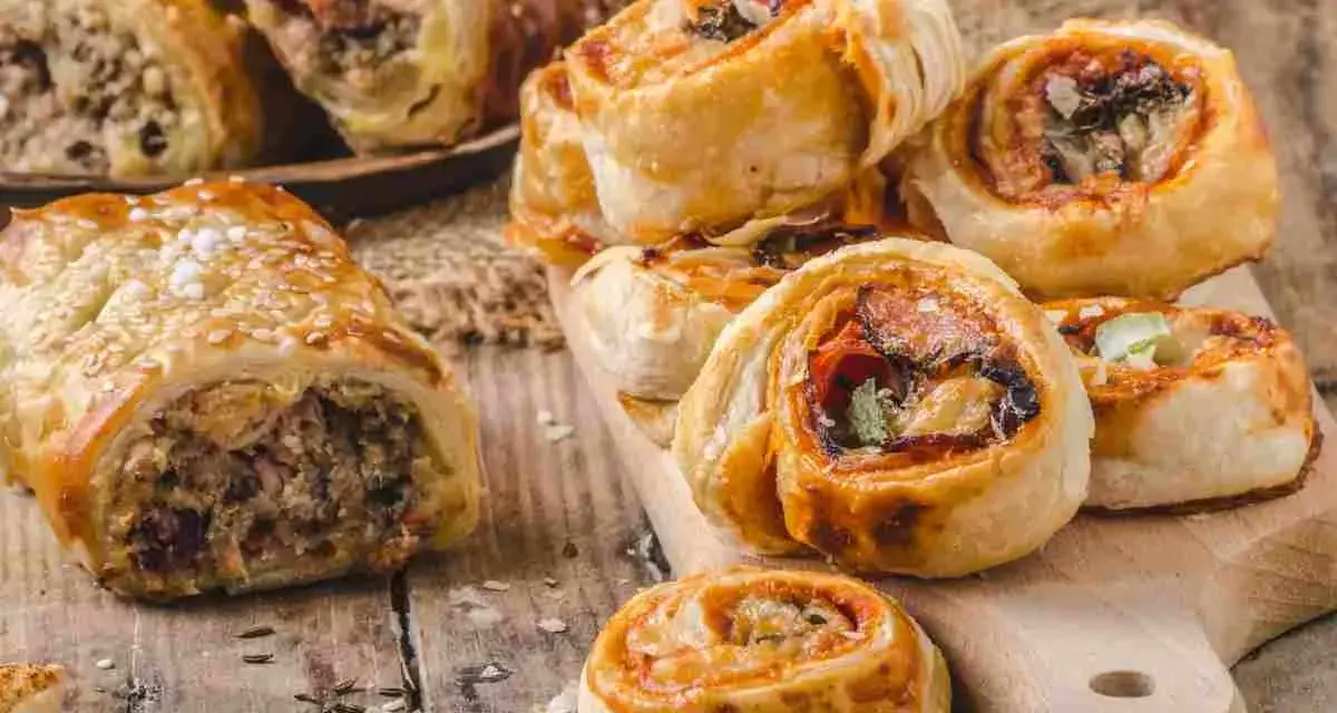 Rustici con pasta sfoglia ripieni di carne e verdure, alcuni cosparsi di semi di sesamo, su un tagliere di legno.