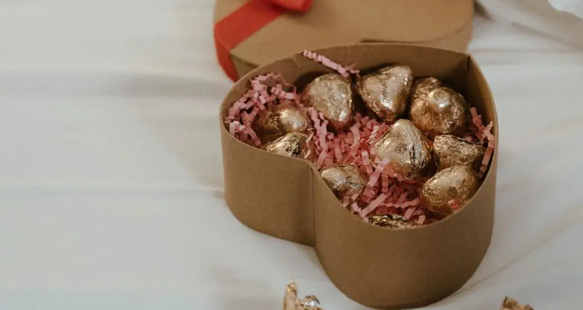 Scatola a forma di cuore con cioccolatini fatti in casa dorati su paglia rosa. Un'altra scatola a forma di cuore con fiocco rosso è sullo sfondo.