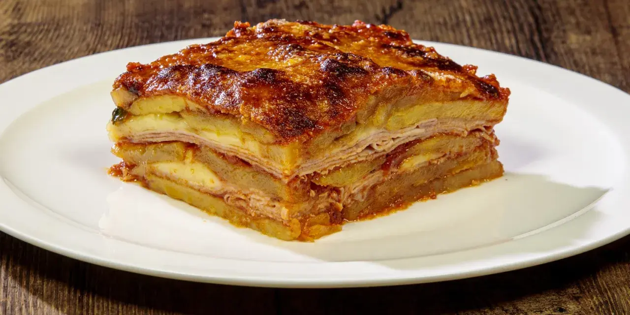 Una fetta di parmigiana di patate, stratificata con formaggio e salumi, servita su un piatto bianco.