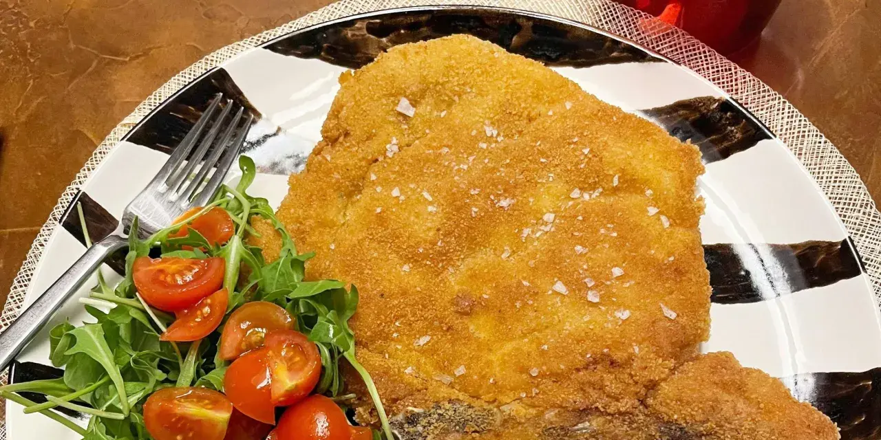 Una dorata cotoletta alla milanese, croccante e cosparsa di sale grosso, servita con un'insalata fresca di rucola e pomodorini.