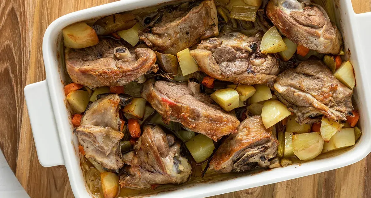 Pezzi succulenti di capretto al forno alla siciliana con patate e carote in una teglia bianca.