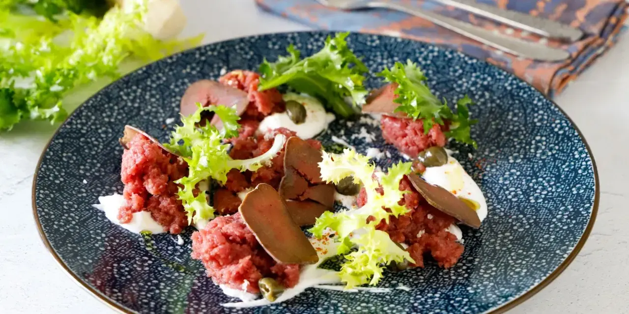 Deliziosa tartare di manzo con lattuga croccante, capperi e una crema bianca su un piatto blu decorato.