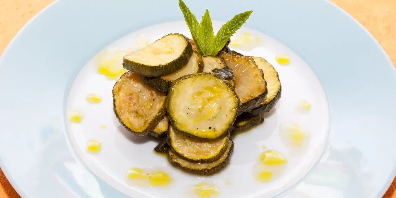 Fette di zucchine alla scapece, condite con olio e guarnite con menta fresca, su un piatto bianco.