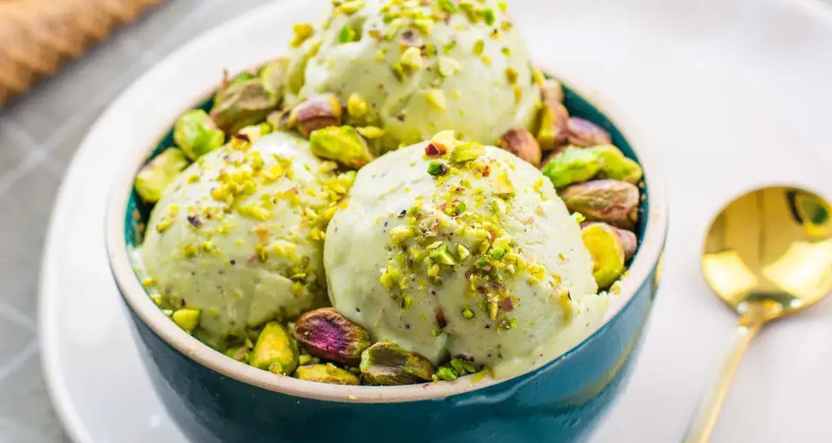 Tre palline di cremoso gelato al pistacchio, guarnite con granella e pistacchi interi, in una ciotola blu.