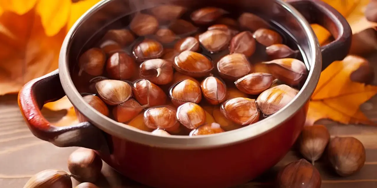 Castagne bollite con buccia in una pentola rossa, circondate da foglie autunnali.