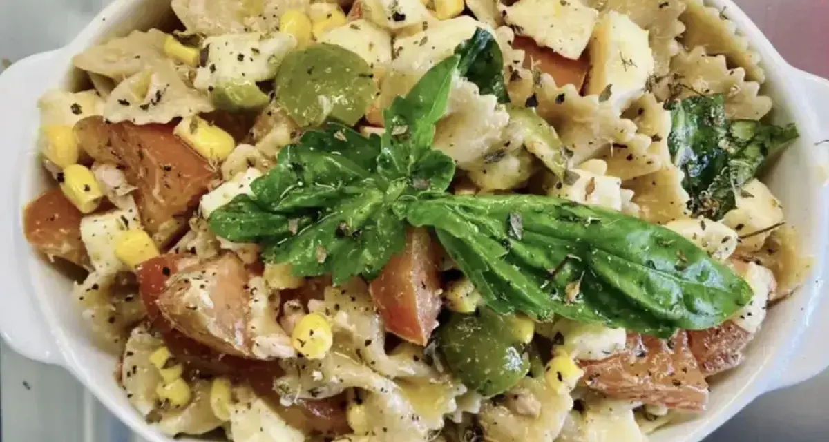 Un'insalata di pasta colorata con farfalle, pomodori, mais, olive verdi e basilico fresco, condita con erbe aromatiche.