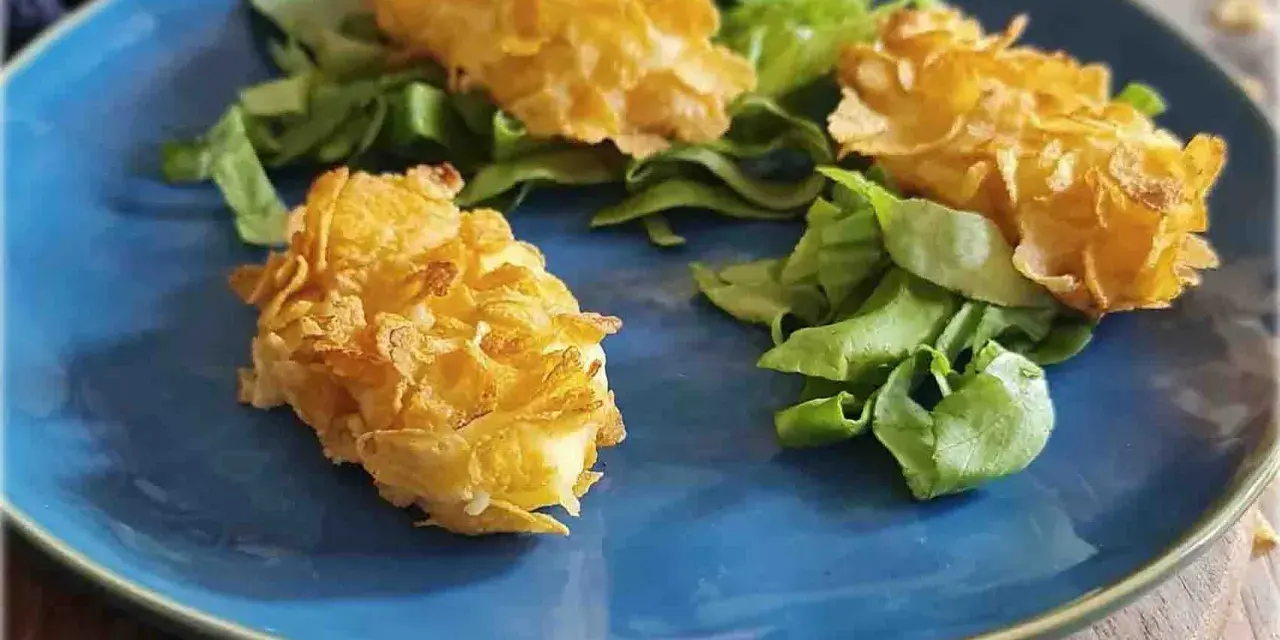 Tre croccanti polpette di patate impanate con fiocchi di mais, servite su un letto di insalata fresca su un piatto blu.