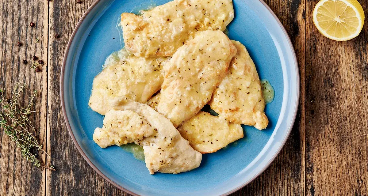 Delizioso petto di pollo in salsa cremosa, guarnito con erbe aromatiche e servito su un piatto blu. Accanto, limone e pepe nero.