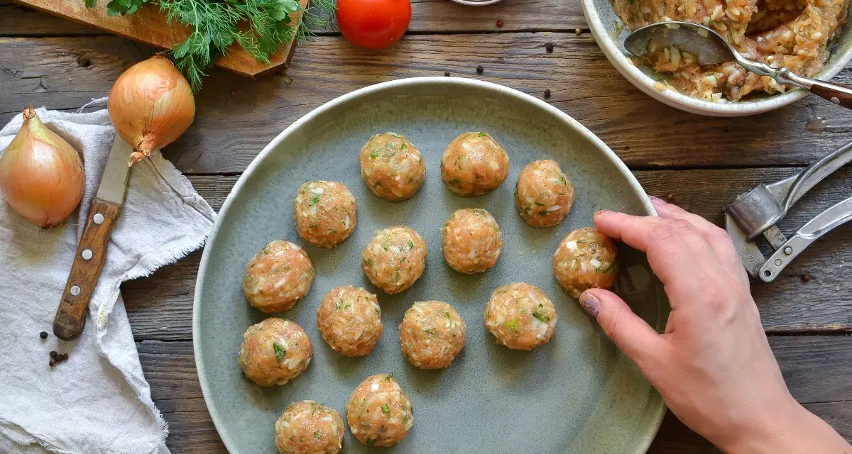 Polpette di pollo pronte per essere cucinate, disposte su un piatto grigio, con ingredienti freschi come cipolle e pomodoro sullo sfondo.