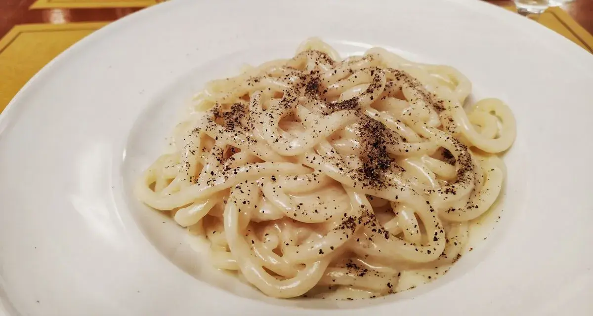 Pici cacio e pepe, un classico piatto romano, servito in un piatto bianco, con abbondante pepe nero macinato.