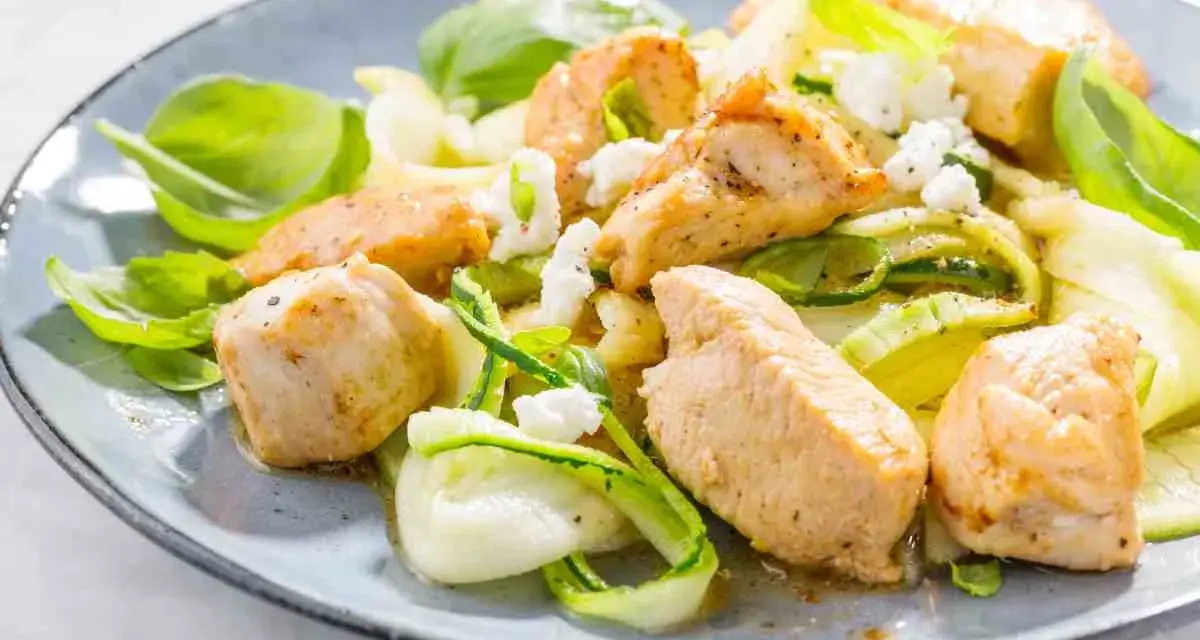 Succulento pollo e zucchine a spirale, guarnito con formaggio sbriciolato e basilico fresco, servito su un piatto blu.