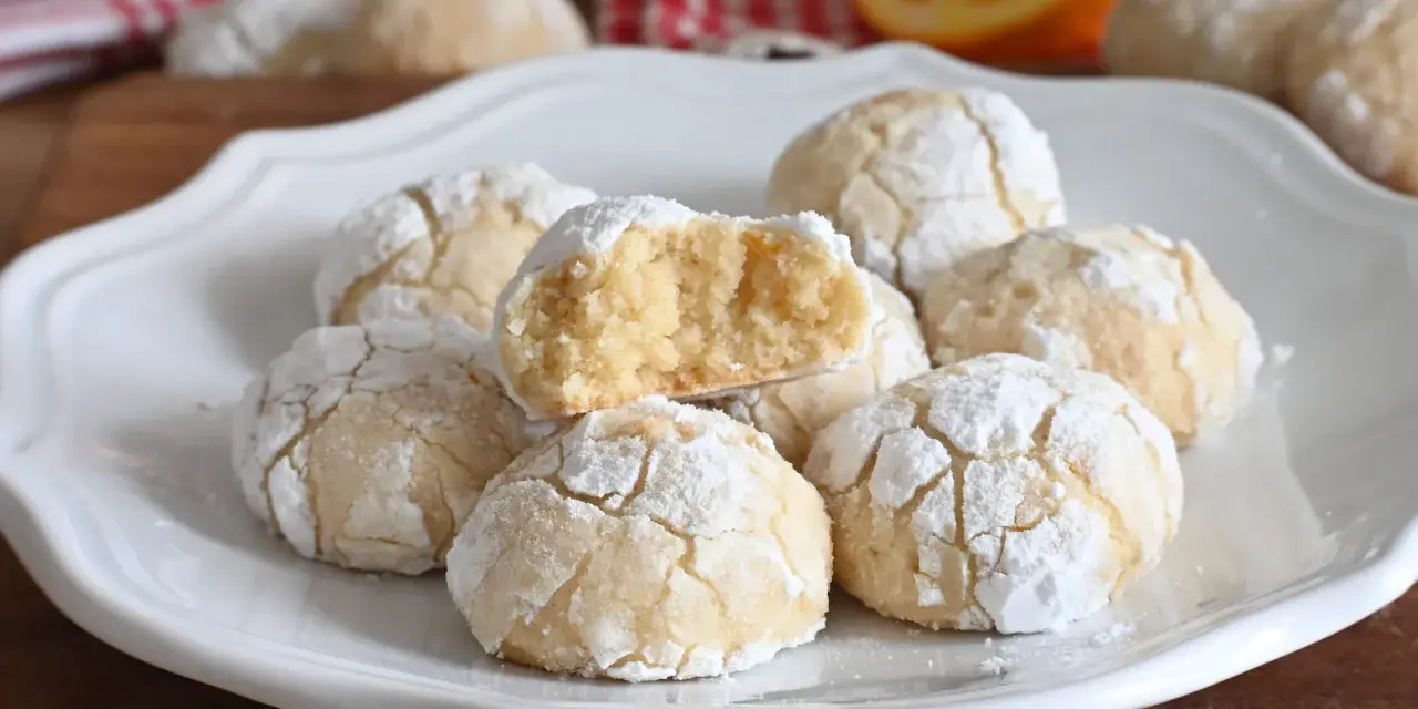Biscotti alla ricotta soffici e profumati, cosparsi di zucchero a velo, con una fetta d'arancia sullo sfondo.