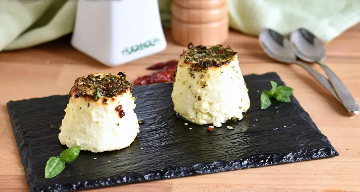 Due tortini di ricotta al forno, dorati e profumati, serviti su ardesia con basilico e peperoncino.
