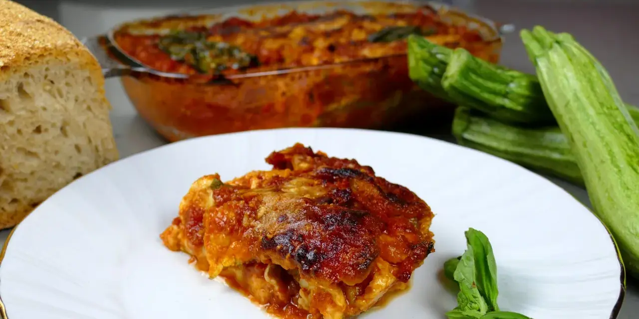 Una fetta di parmigiana di zucchine alla siciliana, dorata e succulenta, accanto a zucchine fresche e pane casereccio.