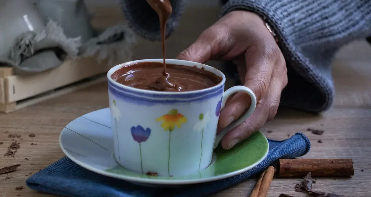 Una mano versa cioccolata fatta in casa in una tazza fiorata. Profumo di cannella e scaglie di cioccolato sul tavolo.