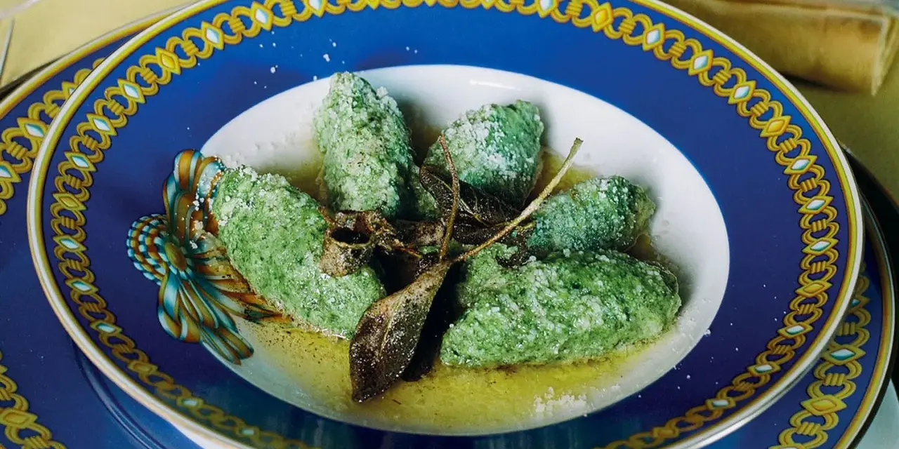 Strangolapreti alla trentina, verdi e saporiti, serviti in un piatto blu decorato con burro fuso e parmigiano.
