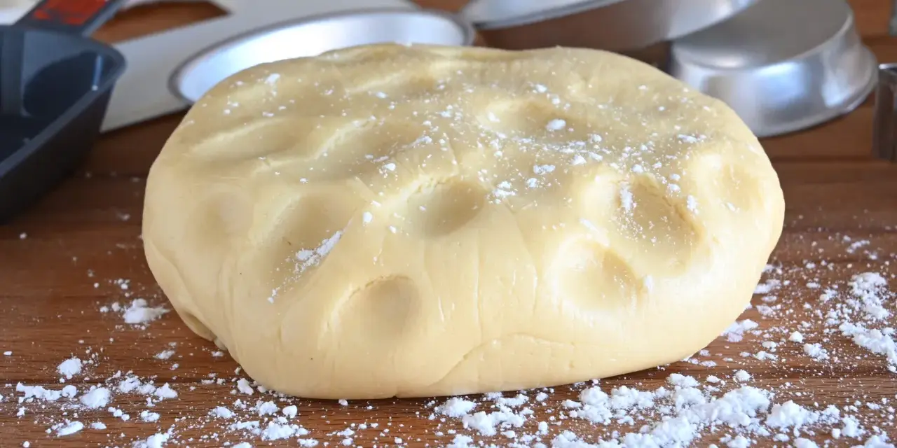 Impasto di pasta frolla con olio, pronto per essere lavorato. Si intravedono stampi per torte e utensili da cucina.