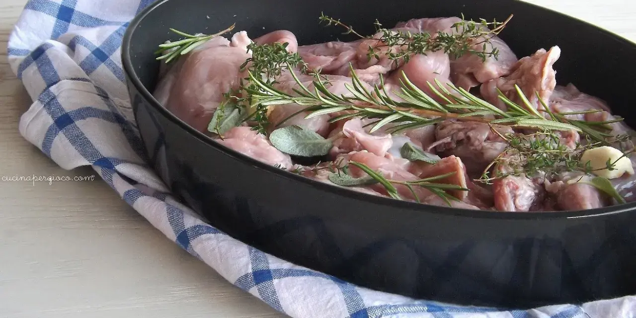 Pezzi di coniglio crudo in una teglia, pronti per essere cucinati al forno con rosmarino, salvia e aglio.