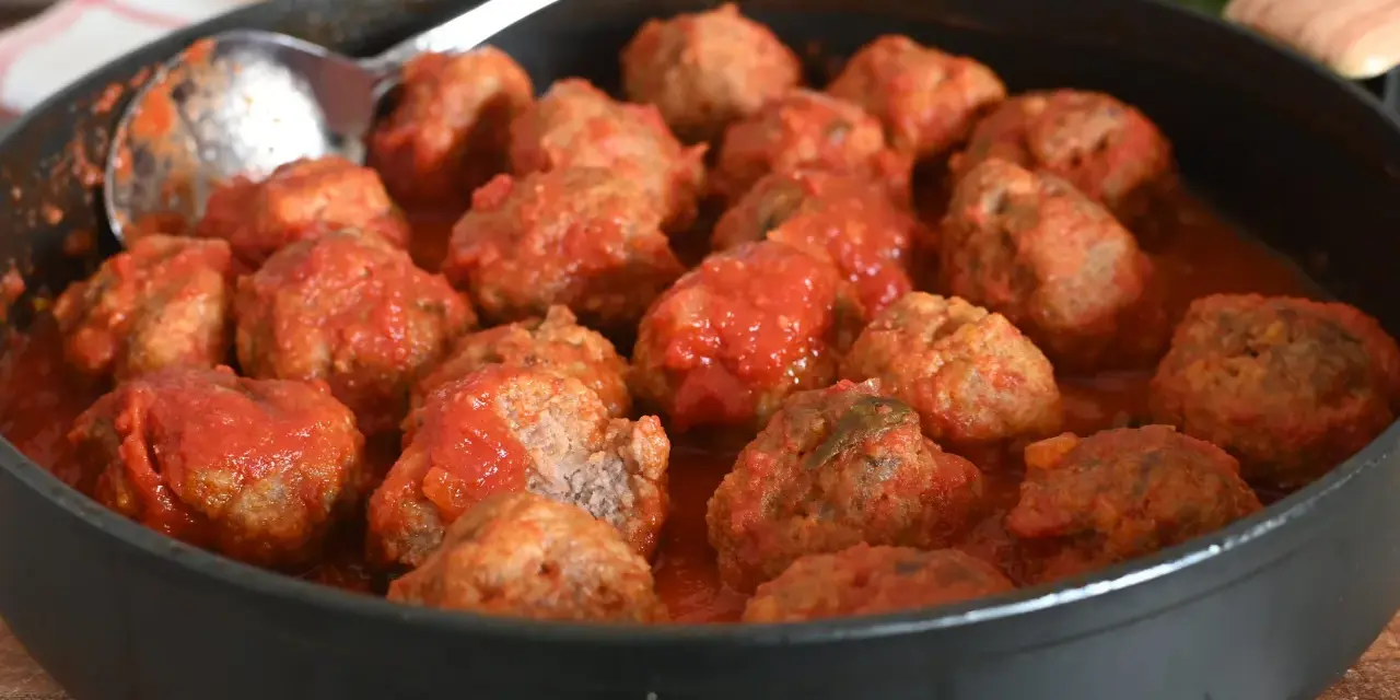 Un tegame pieno di polpette al sugo, pronte per essere gustate. Un cucchiaio d'argento è appoggiato sul bordo, invitando a servirle.