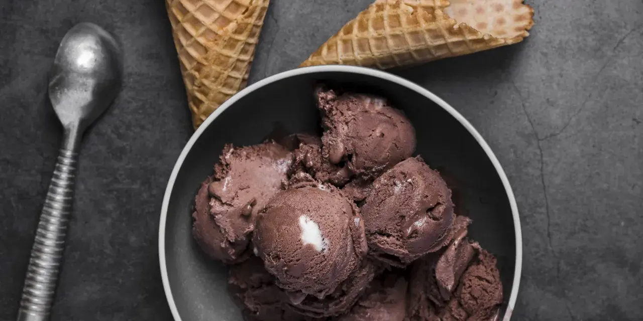Coppa di **gelato al cioccolato** cremosa, con coni vuoti e un cucchiaio d'argento su sfondo scuro.