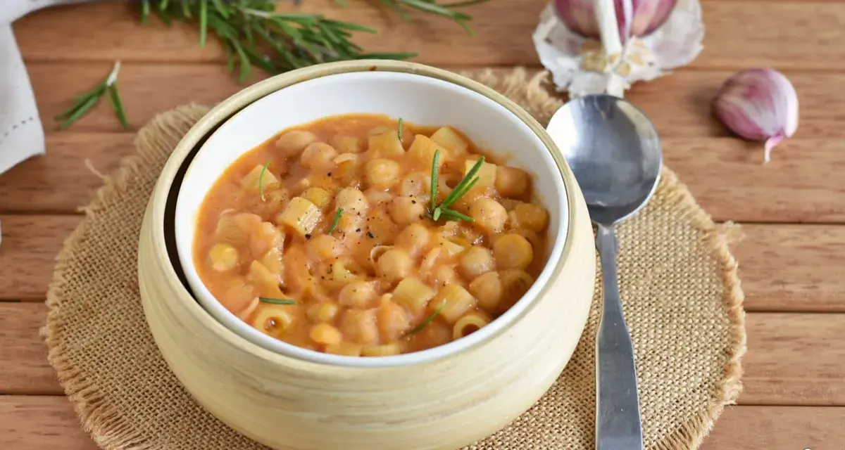 Un piatto fumante di pasta e ceci, guarnito con rosmarino fresco, su un tavolo di legno. Accanto, spicchi d'aglio e un cucchiaio.