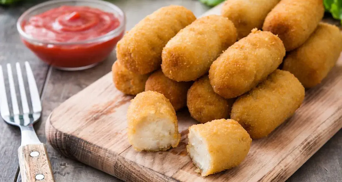 Crocchette di patate dorate e croccanti, servite su un tagliere di legno con ketchup e una forchetta.