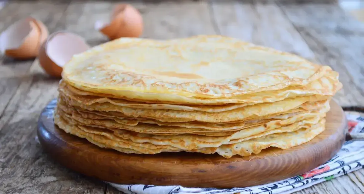 Pila di crepes dorate, perfette per una crepes ricetta senza burro. Guscio d'uovo sullo sfondo.