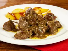 Un saporito abbacchio alla cacciatora, tenero e succulento, servito con patate arrosto su un piatto bianco.
