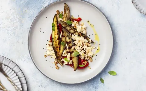 Deliziosa insalata di melanzane grigliate con feta sbriciolata, pomodoro, noci e semi di sesamo nero, condita con olio d'oliva.