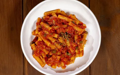 Un piatto di pasta alla pizzaiola, con rigatoni conditi da un sugo rosso ricco di pomodoro, capperi e origano, servito in una ciotola bianca su un tavolo di legno.