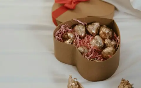 Scatola a forma di cuore con cioccolatini fatti in casa dorati su paglia rosa. Un'altra scatola a forma di cuore con fiocco rosso è sullo sfondo.