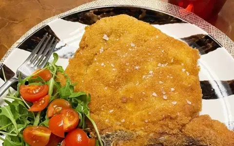 Una dorata cotoletta alla milanese, croccante e cosparsa di sale grosso, servita con un'insalata fresca di rucola e pomodorini.