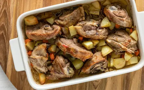 Pezzi succulenti di capretto al forno alla siciliana con patate e carote in una teglia bianca.