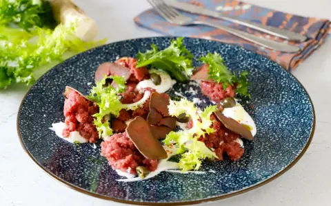 Deliziosa tartare di manzo con lattuga croccante, capperi e una crema bianca su un piatto blu decorato.