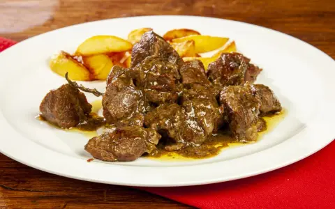 Un saporito abbacchio alla cacciatora, tenero e succulento, servito con patate arrosto su un piatto bianco.