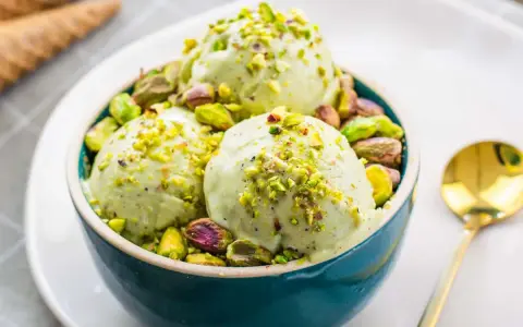 Tre palline di cremoso gelato al pistacchio, guarnite con granella e pistacchi interi, in una ciotola blu.