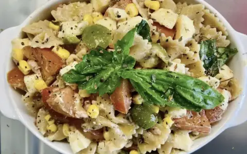 Un'insalata di pasta colorata con farfalle, pomodori, mais, olive verdi e basilico fresco, condita con erbe aromatiche.