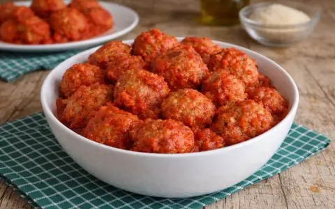 Un primo piatto invitante: polpette di pane al sugo, morbide e saporite, servite in una ciotola bianca su un tovagliolo a quadretti verdi.