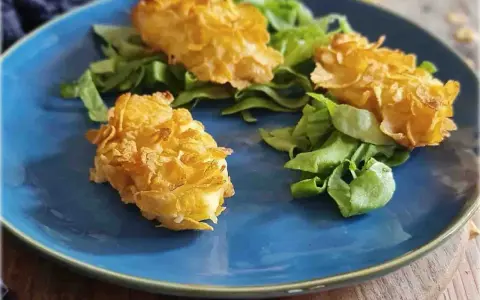 Tre croccanti polpette di patate impanate con fiocchi di mais, servite su un letto di insalata fresca su un piatto blu.