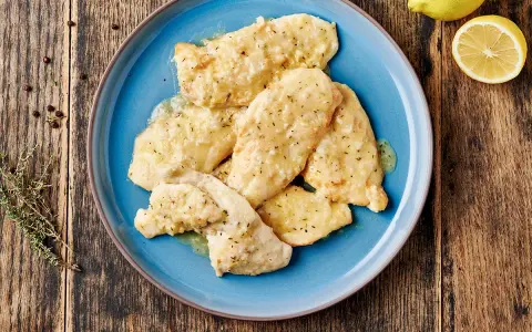 Delizioso petto di pollo in salsa cremosa, guarnito con erbe aromatiche e servito su un piatto blu. Accanto, limone e pepe nero.