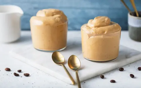 Due bicchieri di cremosa schiuma di caffè, perfetti per una ricetta crema di caffè senza panna. Due cucchiaini dorati e chicchi di caffè completano la scena.